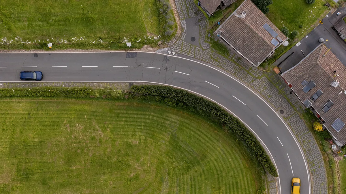 Bovenaanzicht van een weg die naar rechts buigt, met een hoge heg aan de binnenkant. De eigen auto nadert de bocht van links. Een gestippelde zichtlijn en een maatlijn tonen het deel dat je overziet; voorbij de heg nadert een tegenligger die je niet ziet.